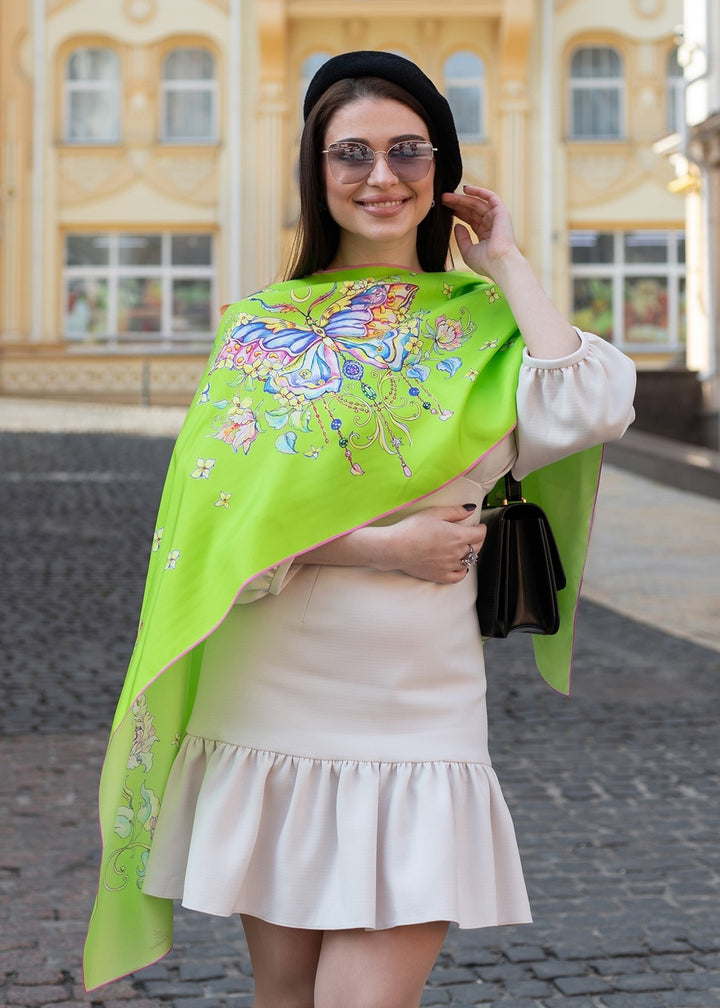 Designer-Seidenschal mit Schmetterlingsmotiv – grüne Seidenschal mit farbenfrohem Schmetterling und Blüten, über die Schultern einer Frau in hellem Kleid getragen, Iryna Feinblatt Design.