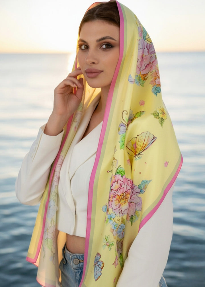 Frau trägt das Seidenkopftuch „Herz aus Blumen in Gelb“ von Iryna Feinblatt über dem Kopf – zarte gelbe Seide mit Blumenmotiv im Morgenlicht am Meer